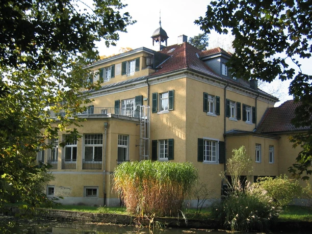 Haus Eulenbroich, Rückansicht Gelbes Herrenhaus mit roten Dachziegeln, umgeben von Bäumen und Schilf, bei sonnigem Wetter.
