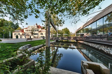 Schloss Eulenbroich Herrenhaus mit modernem Glasanbau, umgeben von grünem Park und reflektierendem Teich im Vordergrund.
