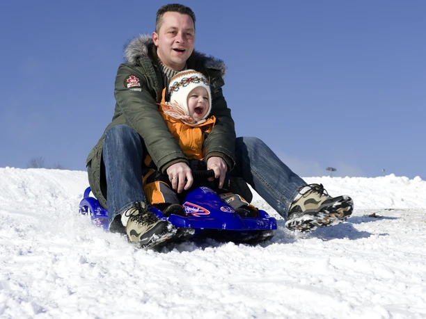 Rodelspaß Mann und Kind rodeln fröhlich im Schnee auf einem blauen Schlitten unter klarem, blauem Himmel.