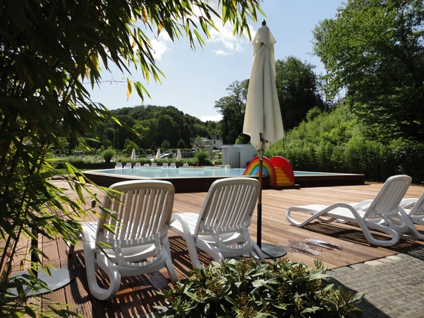 Sonnenliegen Ein sonniger Holzsteg mit weißen Liegestühlen, Sonnenschirm und Pool in grüner, bewaldeter Umgebung.