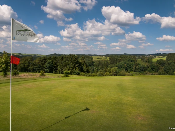 Golfplatz Golfplatz mit Flaggen vor hügeliger Landschaft, bedeckt von Wiesen und Wäldern unter blauem Himmel.