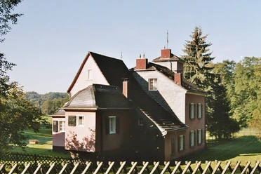 Haus Stade, Ansicht von Lüghauser Straße Rosa zweistöckiges Haus mit steilem Dach und Schornsteinen, umgeben von Bäumen und grünem Rasen.