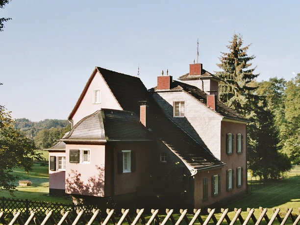 Haus Stade, Ansicht von Lüghauser Straße Rosa zweistöckiges Haus mit steilem Dach und Schornsteinen, umgeben von Bäumen und grünem Rasen.