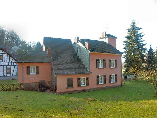 Haus Stade, Ansicht von Lüghauser Straße Ein rosafarbenes, zweistöckiges Haus mit geschlossenen Fensterläden und einem großen Garten im Vordergrund.