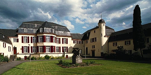Haus Venauen, Vorderansicht Historisches Gebäude mit Schindeldach, weißer Fassade, Garten mit Statue im Vordergrund.
