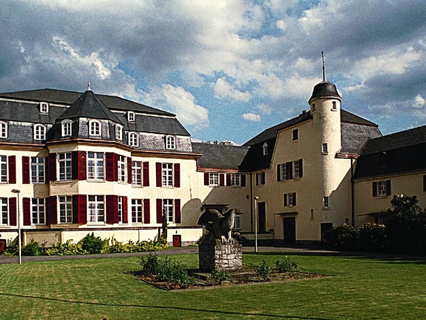 Haus Venauen, Vorderansicht Historisches Gebäude mit Schindeldach, weißer Fassade, Garten mit Statue im Vordergrund.