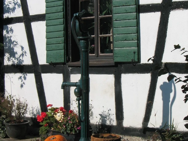 Siedlung Hofferhof, Detail Fachwerkhaus mit grünen Fensterläden, historische Wasserpumpe, Topfpflanzen und Kürbis im Vordergrund.