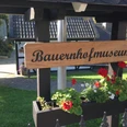 Bauernhofmuseum Holzschild des Bauernhofmuseums vor einem traditionellen Gebäude mit blühenden Geranien.