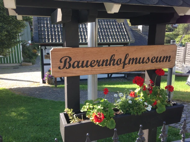 Bauernhofmuseum Holzschild des Bauernhofmuseums vor einem traditionellen Gebäude mit blühenden Geranien.