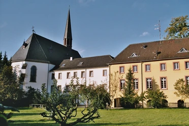 Pfarrkirche und ehem. Kloster, Rückansicht Kirche mit spitzem Turm und angeschlossenen Gebäuden, umgeben von grüner Wiese und Bäumen.
