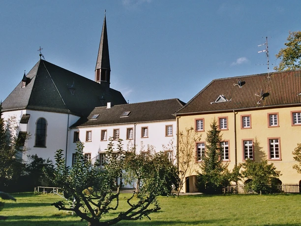 Pfarrkirche und ehem. Kloster, Rückansicht Kirche mit spitzem Turm und angeschlossenen Gebäuden, umgeben von grüner Wiese und Bäumen.