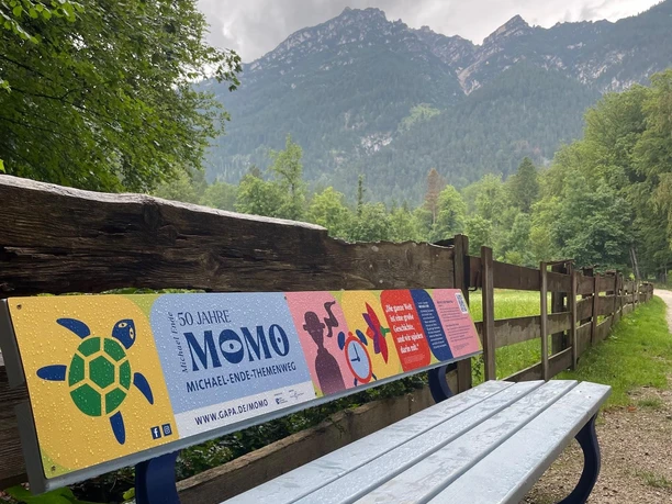 Michael-Ende-Bank Du siehst eine bunte Bank am Weg. Hinter der Bank stehen hohe Berge unter grauem Himmel.