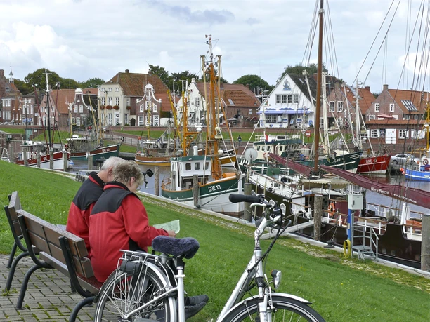 Zwei Radfahrende auf einer Bank am Hafen Greetsiel mit Blick auf Fischerboote und Backsteinhäuser.