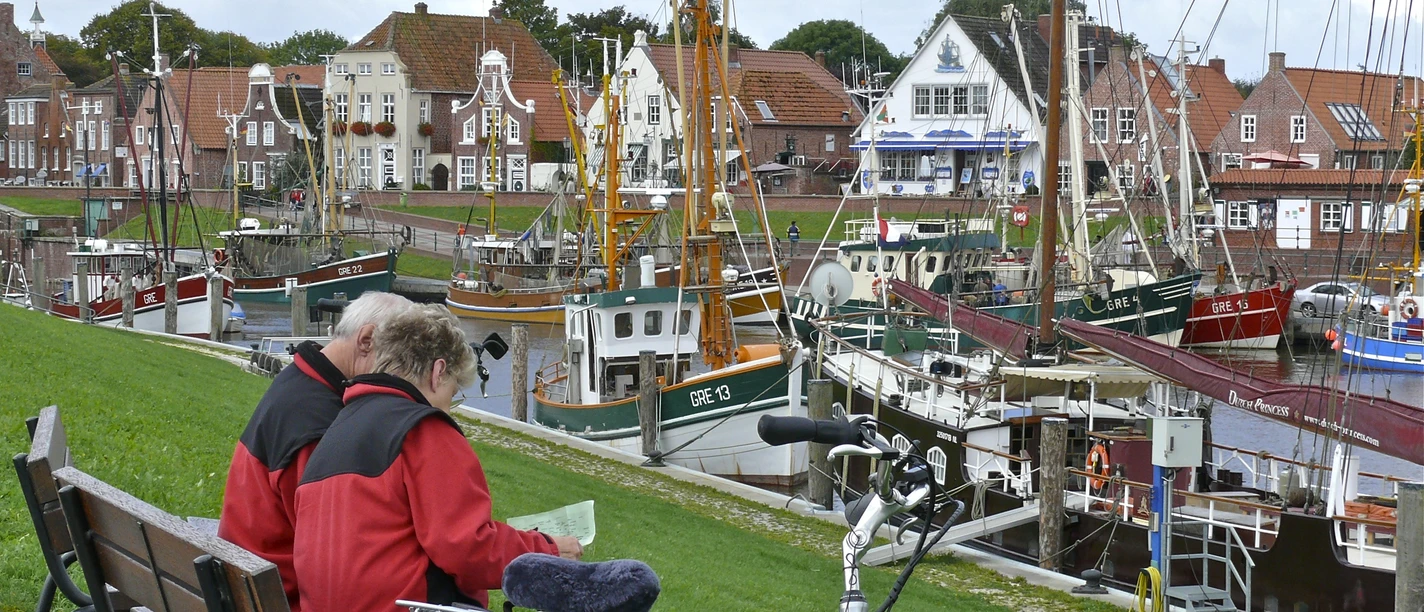 Hafen Greetsiel Radfahrer Zwei Radfahrende auf einer Bank am Hafen Greetsiel mit Blick auf Fischerboote und Backsteinhäuser.