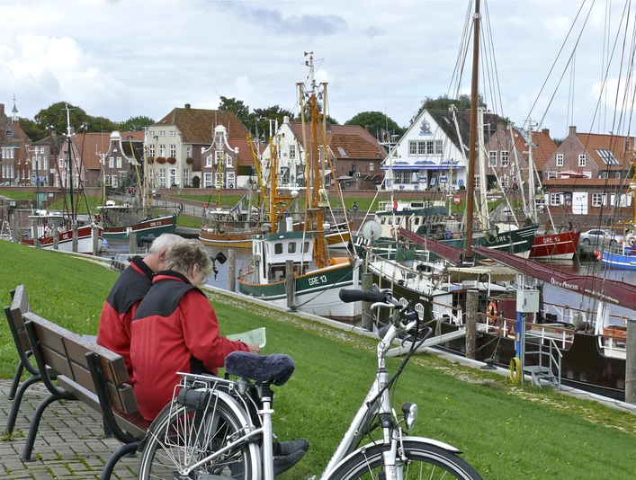 Hafen Greetsiel Radfahrer Zwei Radfahrende auf einer Bank am Hafen Greetsiel mit Blick auf Fischerboote und Backsteinhäuser.