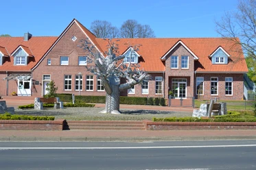 Bürgerbaum Dersum Metallskulptur eines stilisierten Baumes vor rotem Backsteingebäude mit Satteldach bei blauem Himmel.