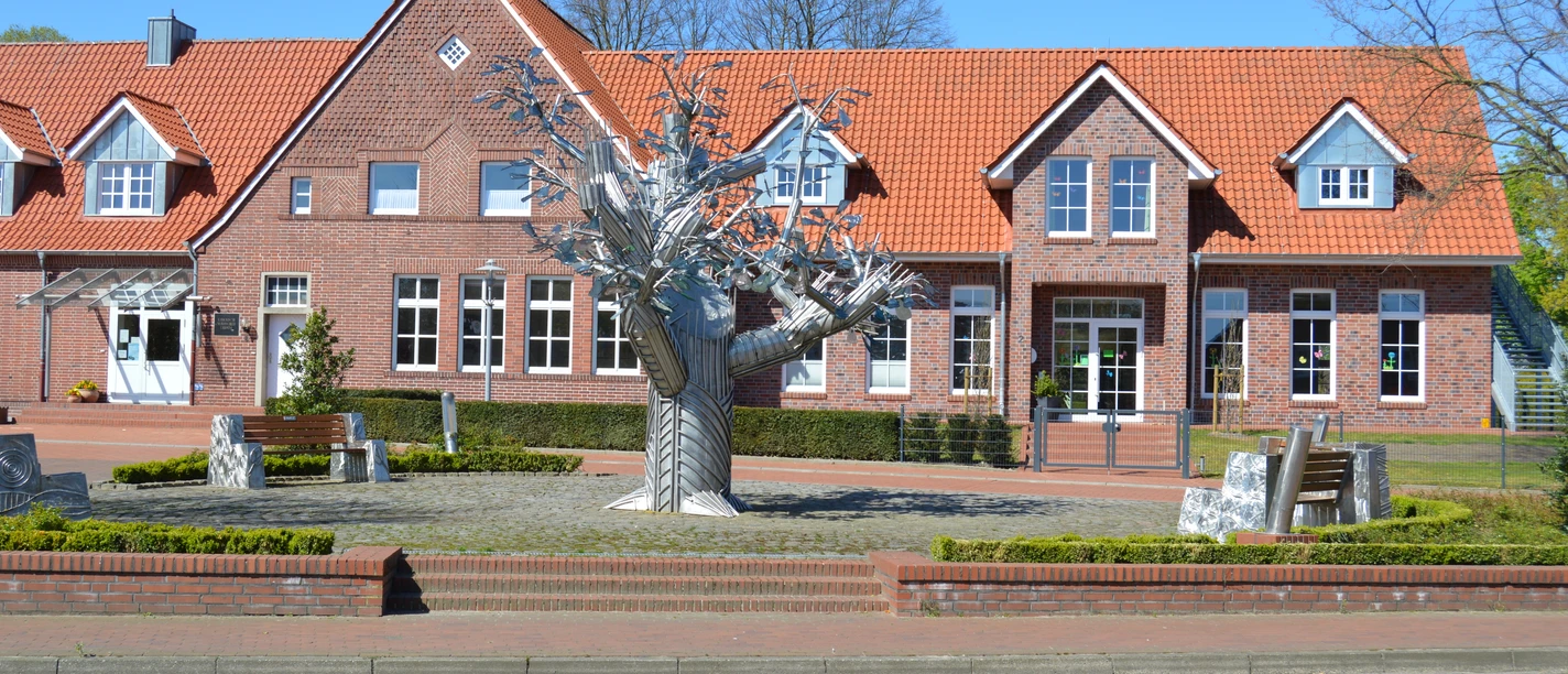 Bürgerbaum Dersum Metallskulptur eines stilisierten Baumes vor rotem Backsteingebäude mit Satteldach bei blauem Himmel.