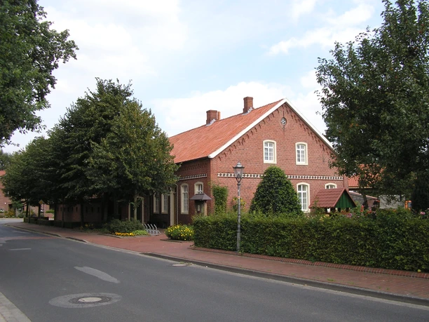 Heimathaus Dörpen Backsteingebäude mit rotem Ziegeldach, Hecken, Bäumen und Laternen an einer ruhigen Straße.