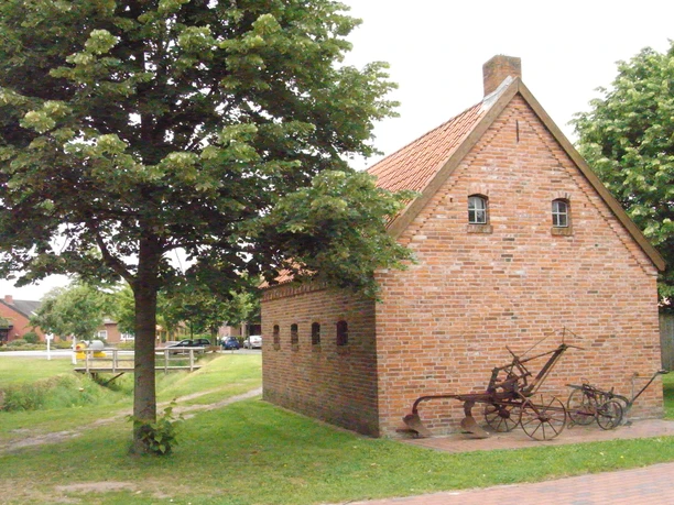 Heimathaus Dörpen Backsteingebäude mit rotem Ziegeldach und alten landwirtschaftlichen Geräten im Freien.