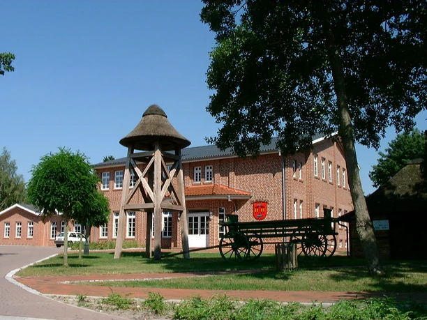 Historischer Dorfplatz Backsteinbau mit Reetdach-Glockenturm und grünem Holzwagen auf einem historischen Dorfplatz.