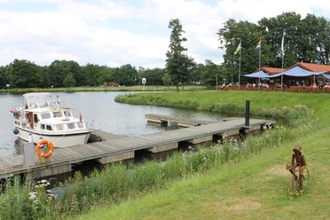 Marinapark Emstal Motorboot an Holzsteg vor einem Seeufer mit Restaurantterrasse und grüner Wiese im Marinapark Emstal