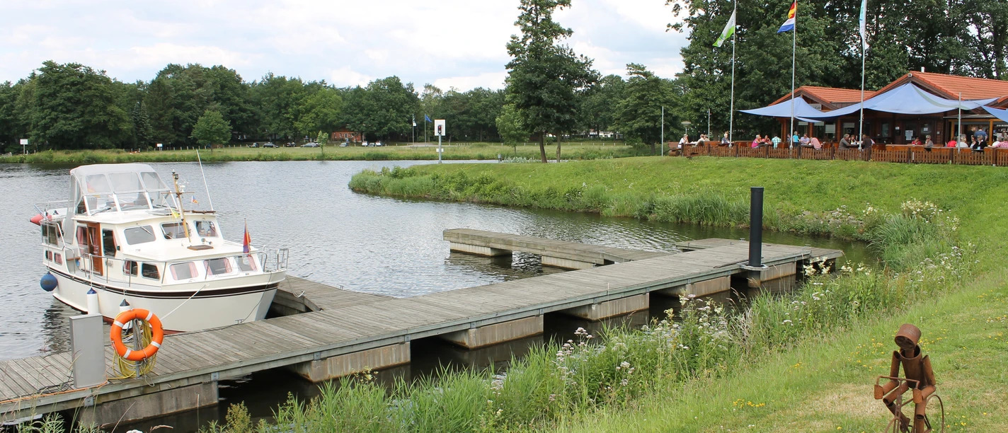 Marinapark Emstal Motorboot an Holzsteg vor einem Seeufer mit Restaurantterrasse und grüner Wiese im Marinapark Emstal