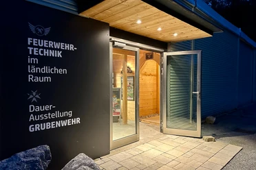 Entrance to the permanent exhibition "Firefighting technology in rural areas" at dusk, with open glass door and illuminated wooden interior.