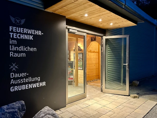 Entrance to the permanent exhibition "Firefighting technology in rural areas" at dusk, with open glass door and illuminated wooden interior.