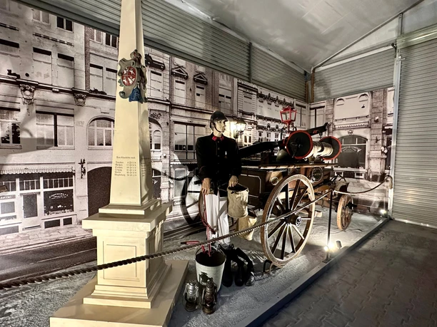 Historical fire department scene with a doll in uniform next to an old fire engine, surrounded by lanterns and an obelisk.