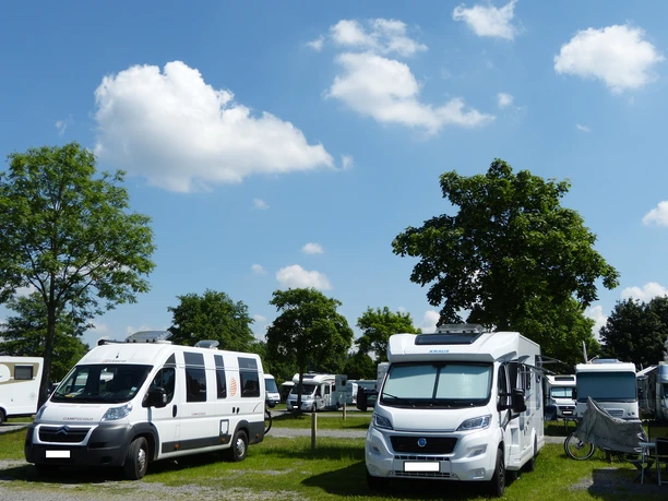 Wohnmobilstellplatz mit mehreren Wohnmobilen unter blauem Himmel und grünen Bäumen an einem sonnigen Tag.