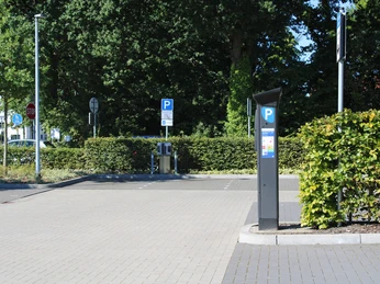 parkplatz-weisse-taube-oldenburger-straße3.jpg Asphaltierter Parkplatz mit markierten Parkplätzen, Parkschein-Automat und grünen Hecken im Hintergrund.