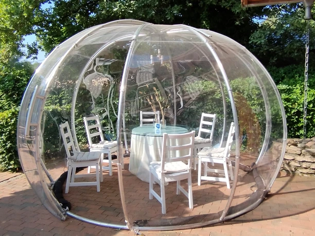 A round glass pavilion with white chairs arranged around a set table surrounded by greenery.