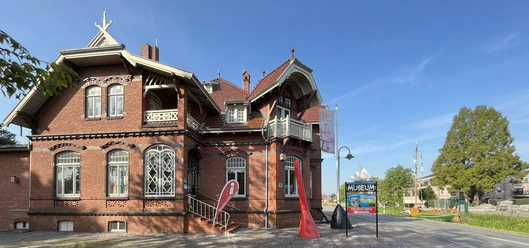 Tourist-Information Rhauderfehn im Fehn- und Schiffahrtsmuseum Westrhauderfehn Historisches rotes Backsteingebäude mit Giebeln und Balkonen, davor eine rote Signalboje.