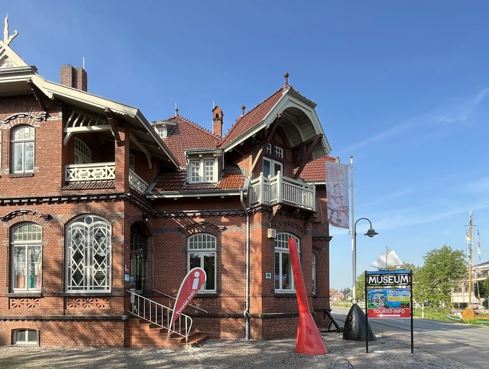 Tourist-Information Rhauderfehn im Fehn- und Schiffahrtsmuseum Westrhauderfehn Backsteingebäude mit Erker und Treppe vor blauem Himmel, davor Fahnen und Schild mit der Aufschrift 'MUSEUM' und 'TOURIST-INFO'