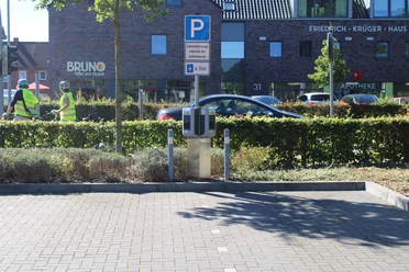 ewe-go-ladestation-oldenburger-straße.jpg Ladestation mit zwei Ladepunkten auf einem Parkplatz, im Hintergrund Gebäude und Bäume.