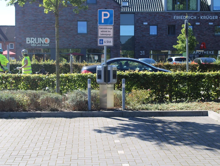 ewe-go-ladestation-oldenburger-straße.jpg Ladestation mit zwei Ladepunkten auf einem Parkplatz, im Hintergrund Gebäude und Bäume.