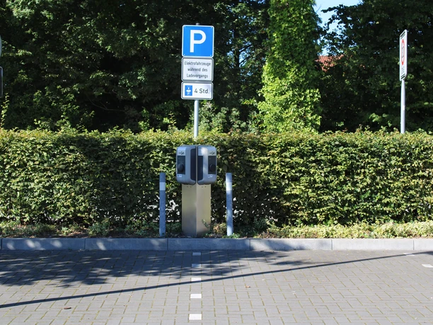 ewe-go-ladestation-oldenburger-straße1.jpg Eine Ladestation für Elektrofahrzeuge steht vor einer grünen Hecke, mitten auf einem Parkplatz, überdacht von Schildern.