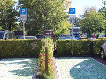 e-ladestation-hotel-am-badepark.jpg Zwei reservierte Parkplätze mit E-Ladestationen vor einem Hotel, umgeben von Hecken und Bäumen.