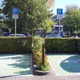 e-ladestation-hotel-am-badepark1.jpg Auf dem Bild sind zwei Parkplätze mit grünen Bodenmarkierungen und einem Schild für E-Ladestationen zu sehen, umgeben von Hecken und Bäumen.