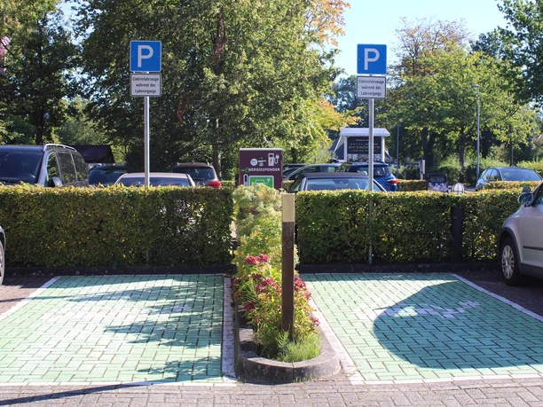 e-ladestation-hotel-am-badepark1.jpg Auf dem Bild sind zwei Parkplätze mit grünen Bodenmarkierungen und einem Schild für E-Ladestationen zu sehen, umgeben von Hecken und Bäumen.