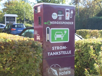 e-ladestation-hotel-am-badepark2.jpg Auf dem Bild ist eine E-Ladestation mit der Aufschrift "ENERGIESPENDER" zu sehen, die von Sträuchern umgeben ist, mit parkenden Autos im Hintergrund.