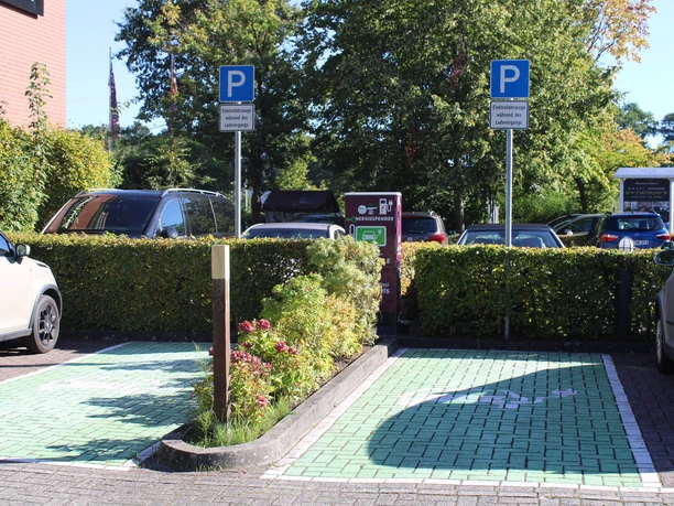 e-ladestation-hotel-am-badepark4.jpg Das Bild zeigt zwei reservierte Parkplätze für Elektrofahrzeuge vor einer E-Ladestation, umgeben von grüner Hecke. Schilder weisen auf Parken während des Ladevorgangs hin.