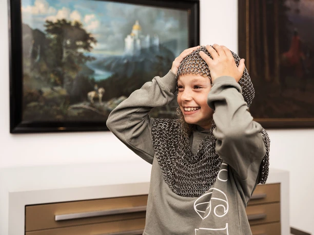 Kinderprogramma | Kreismuseum Wewelsburg Ein Junge lächelt und trägt ein Kettenhemd im Kreismuseum Wewelsburg, im Hintergrund zwei Gemälde.