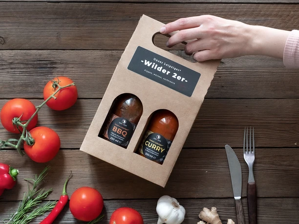 Kern & Stein Saucenbox "Wilder 2er" - Regionale Produkte aus Leipzig Eine Hand hält eine Pappverpackung mit 2 Feinkostsaucen der Kern und Stein Manufaktur aus Gohlis ins Bild, auf dem Tisch darunter liegen Tomaten, Paprika, Knoblauch und Chili, Regionale Produkte aus Leipzig