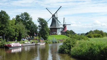 ostfrieslandrundfahrt Zwei historische Windmühlen an einem Kanal, umgeben von grüner Landschaft und blauem Himmel.