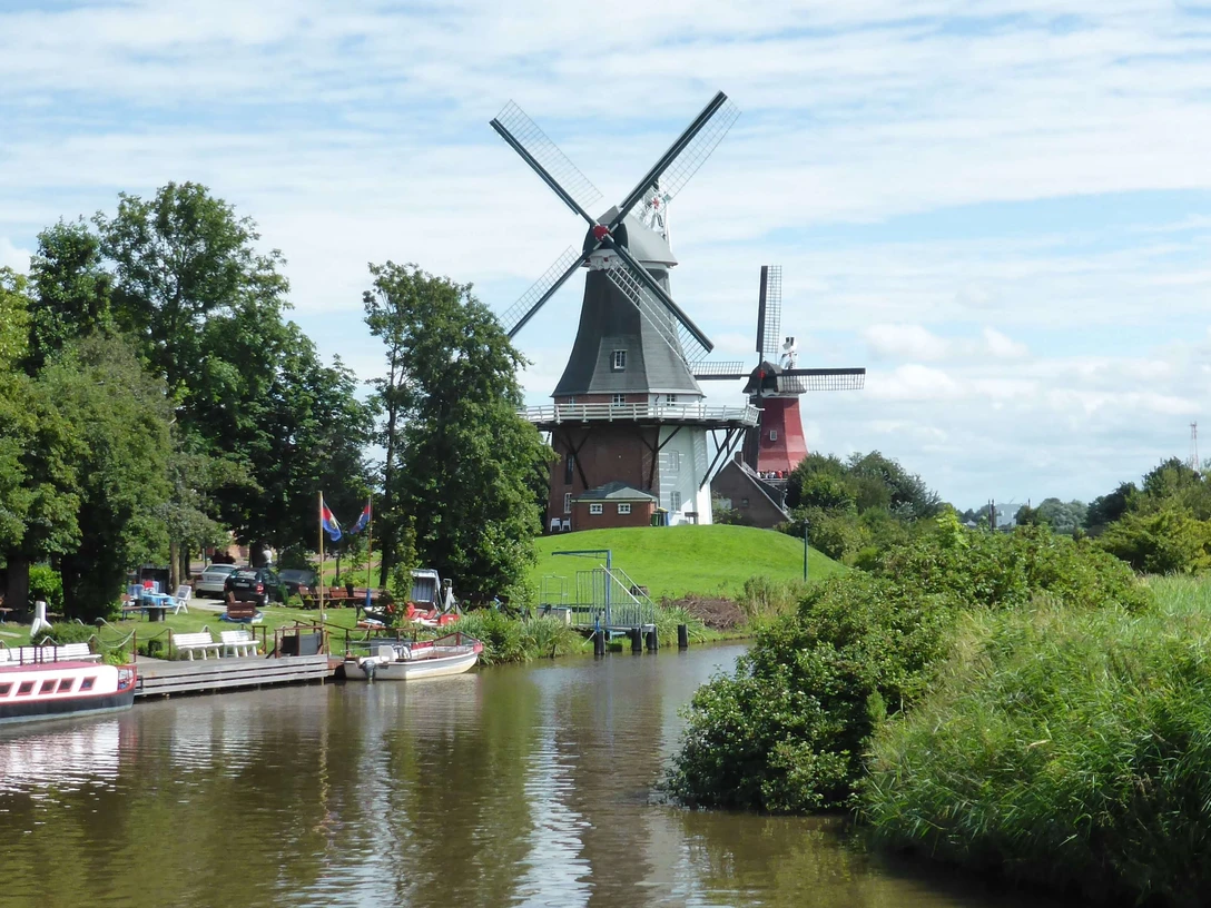 ostfrieslandrundfahrt Zwei historische Windmühlen an einem Kanal, umgeben von grüner Landschaft und blauem Himmel.