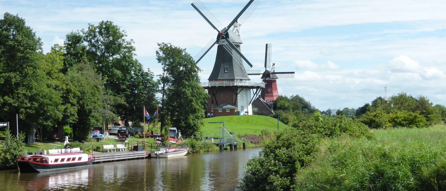 ostfrieslandrundfahrt Zwei historische Windmühlen an einem Kanal, umgeben von grüner Landschaft und blauem Himmel.