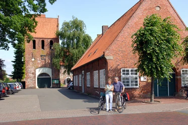 07_056.JPG Ein altes Backsteingebäude mit roten Dachziegeln und Bäumen davor. Zwei Radfahrer stehen lachend daneben.