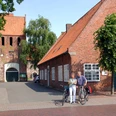 07_056.JPG Ein altes Backsteingebäude mit roten Dachziegeln und Bäumen davor. Zwei Radfahrer stehen lachend daneben.