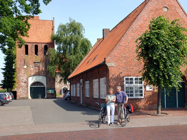 07_056.JPG Ein altes Backsteingebäude mit roten Dachziegeln und Bäumen davor. Zwei Radfahrer stehen lachend daneben.